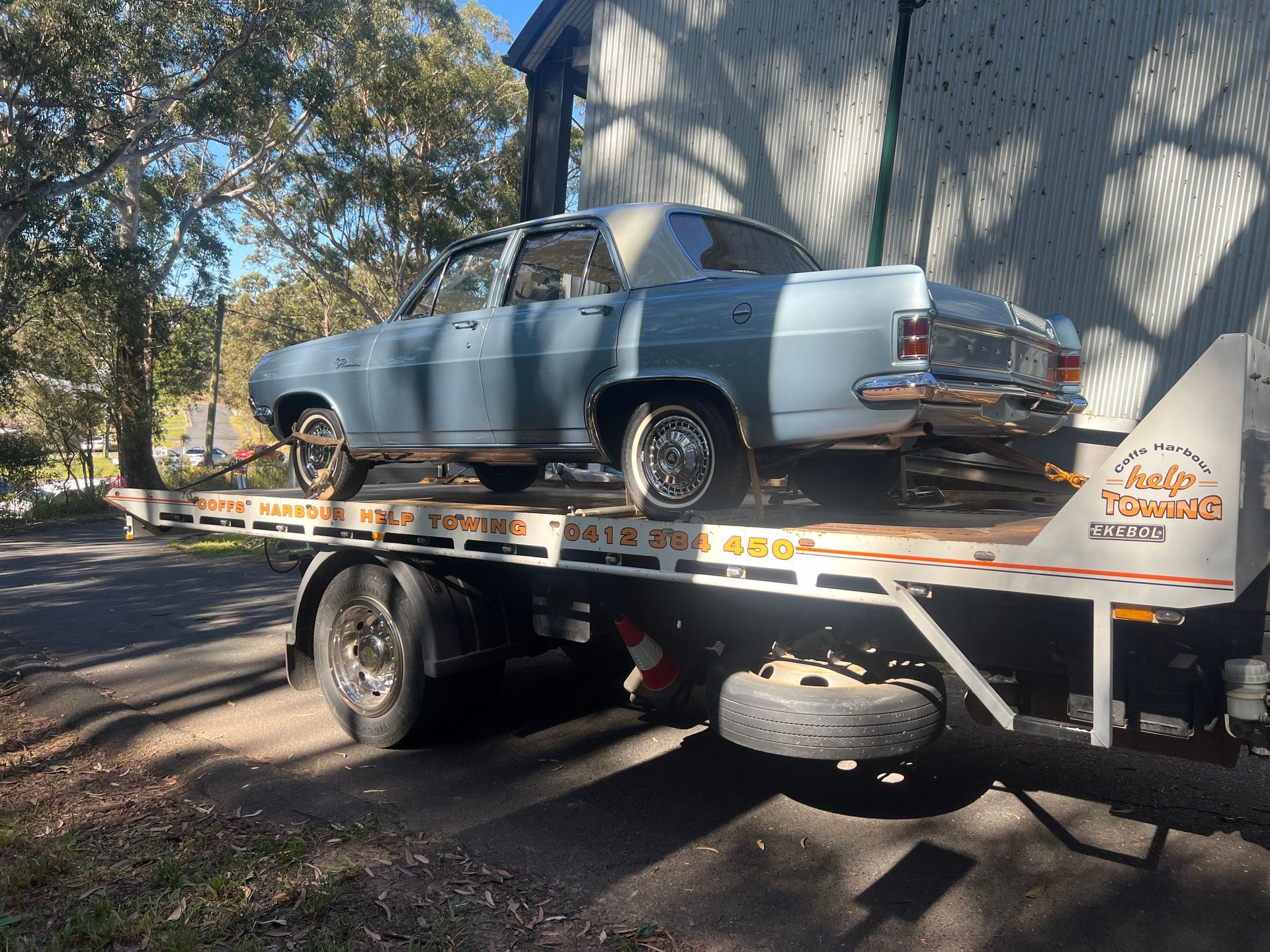 Classic light blue car strapped on a flatbed tow truck under trees.