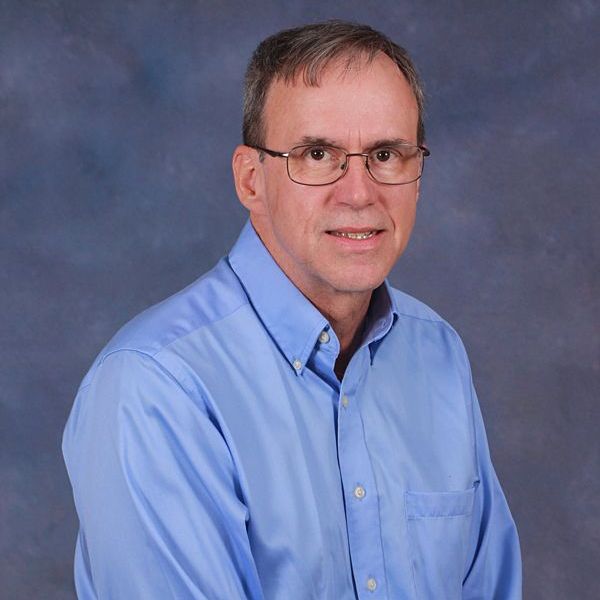 A man wearing glasses and a blue shirt is standing in front of a black background.