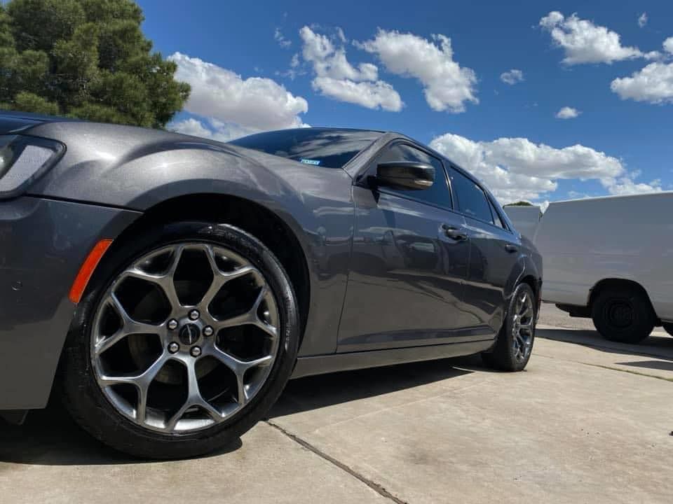 Dark gray sedan parked on a driveway under a blue sky with clouds, low-angle close-up of the front wheel.