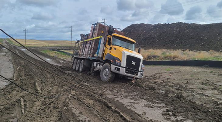 Yellow dump truck stuck in deep mud on a dirt road under a cloudy sky.