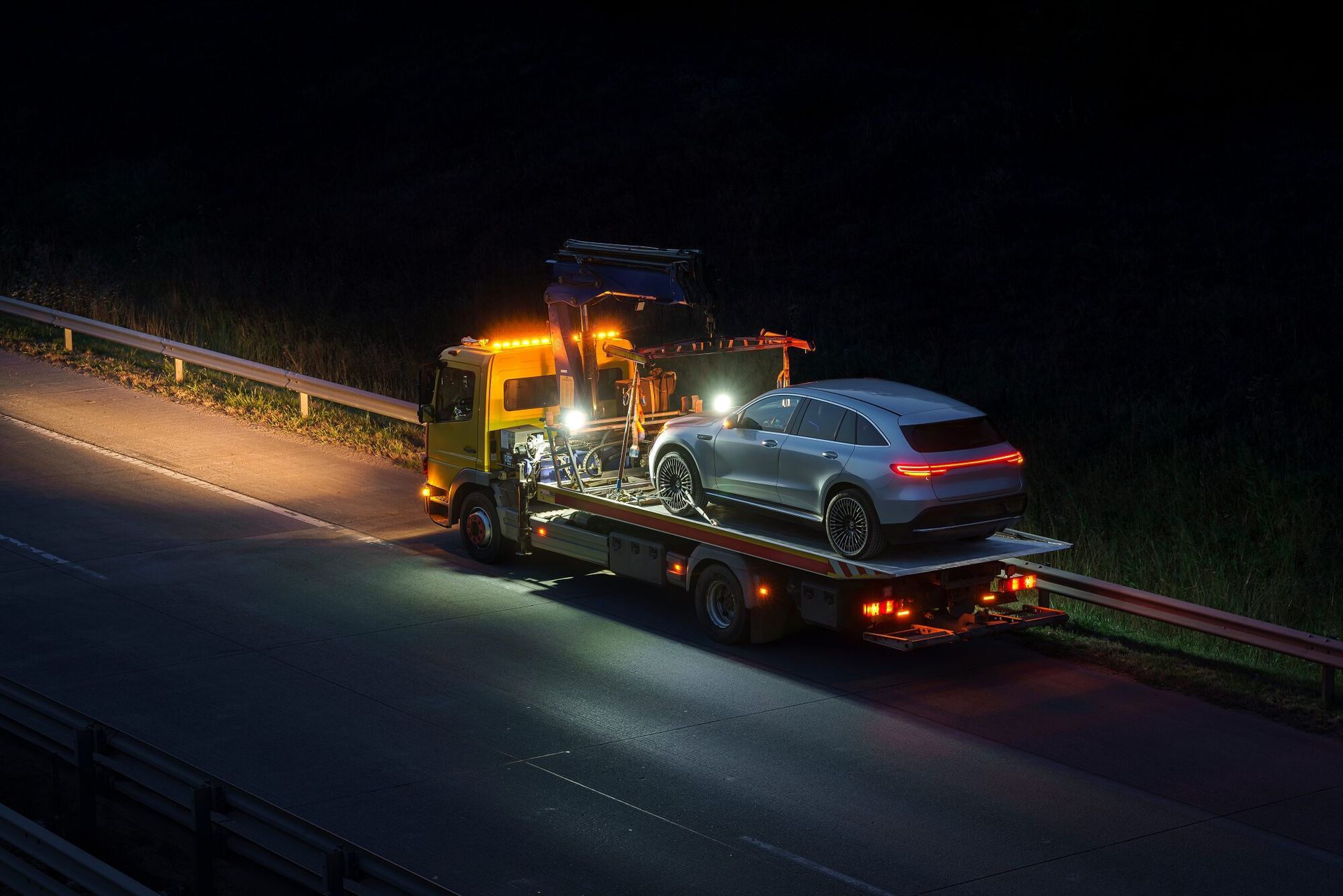 Tow truck with silver car on the bed, illuminated at night on a dark highway.