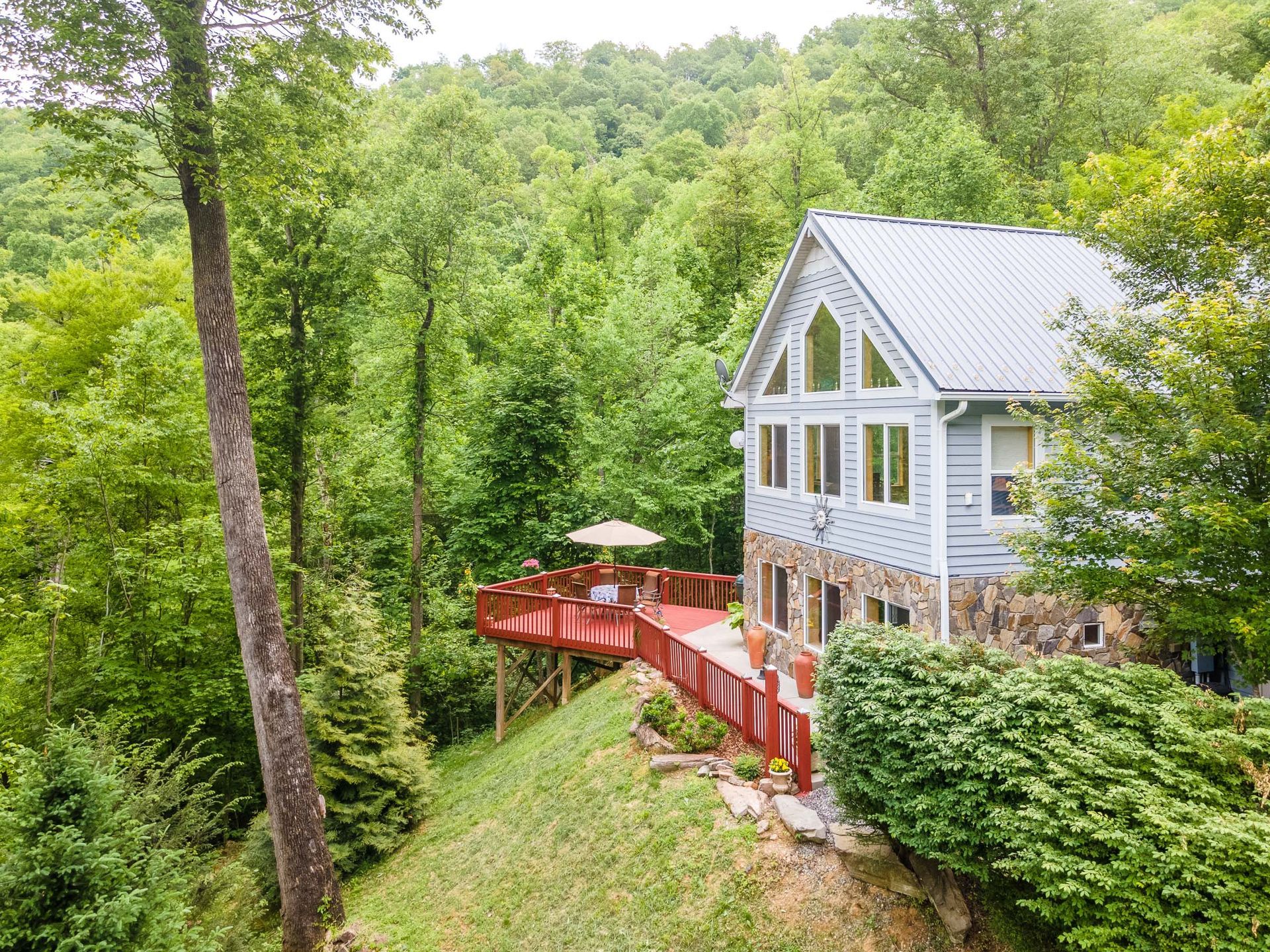 Cottage with red deck nestled in a lush, green forest. Gray siding, stone base, and a white metal roof.