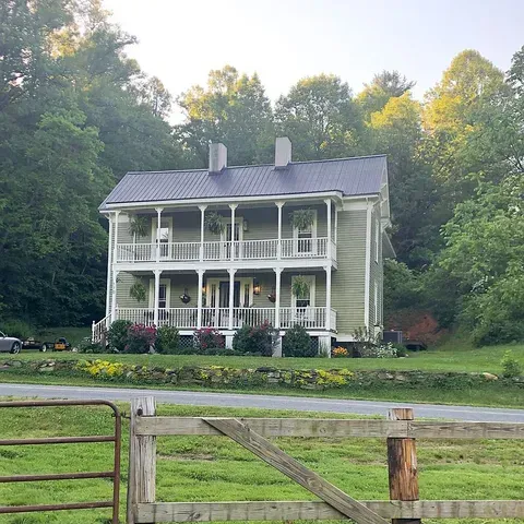 Two-story light green house with double porches. Set in a wooded area, with flowers and a fence in the foreground.
