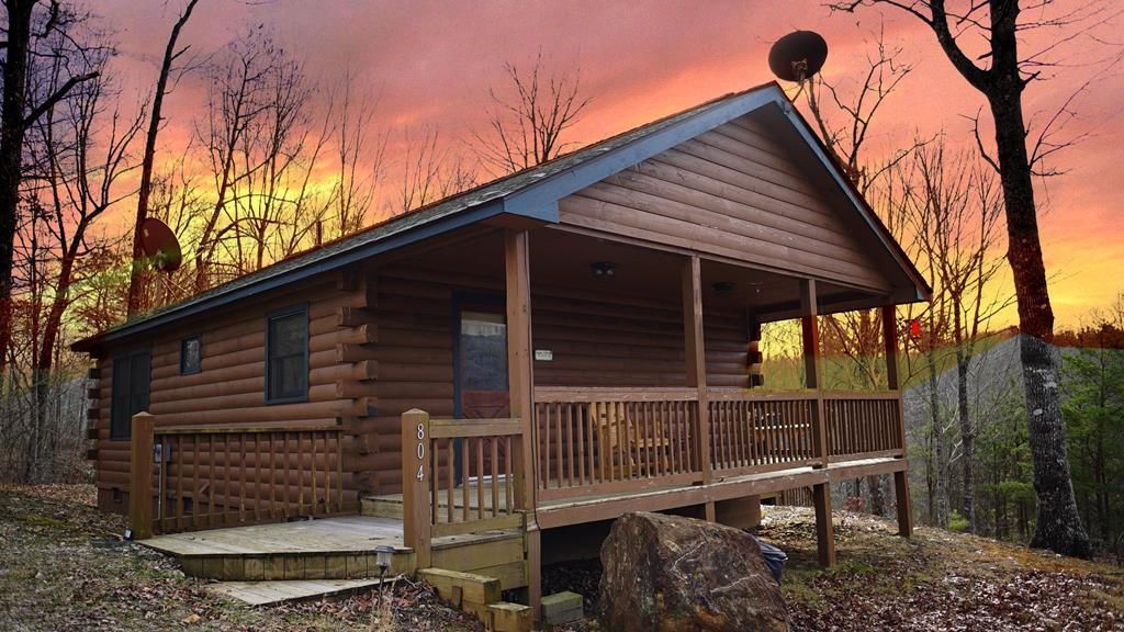 Log cabin with porch and satellite dish, set in woods, with a sunset sky.