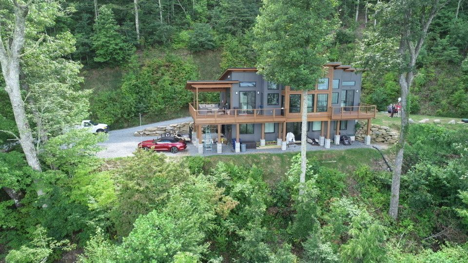 Modern house on a hillside with large deck, surrounded by trees. A red car is parked in the driveway.