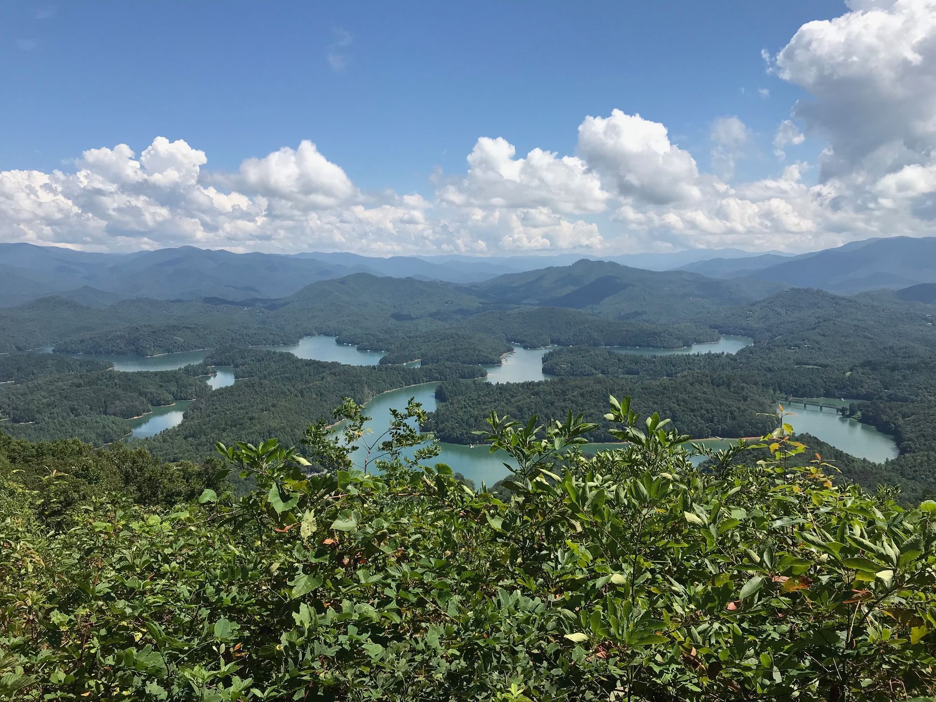 Overhead view of a lush green landscape with multiple lakes nestled amidst forested hills under a cloudy blue sky.