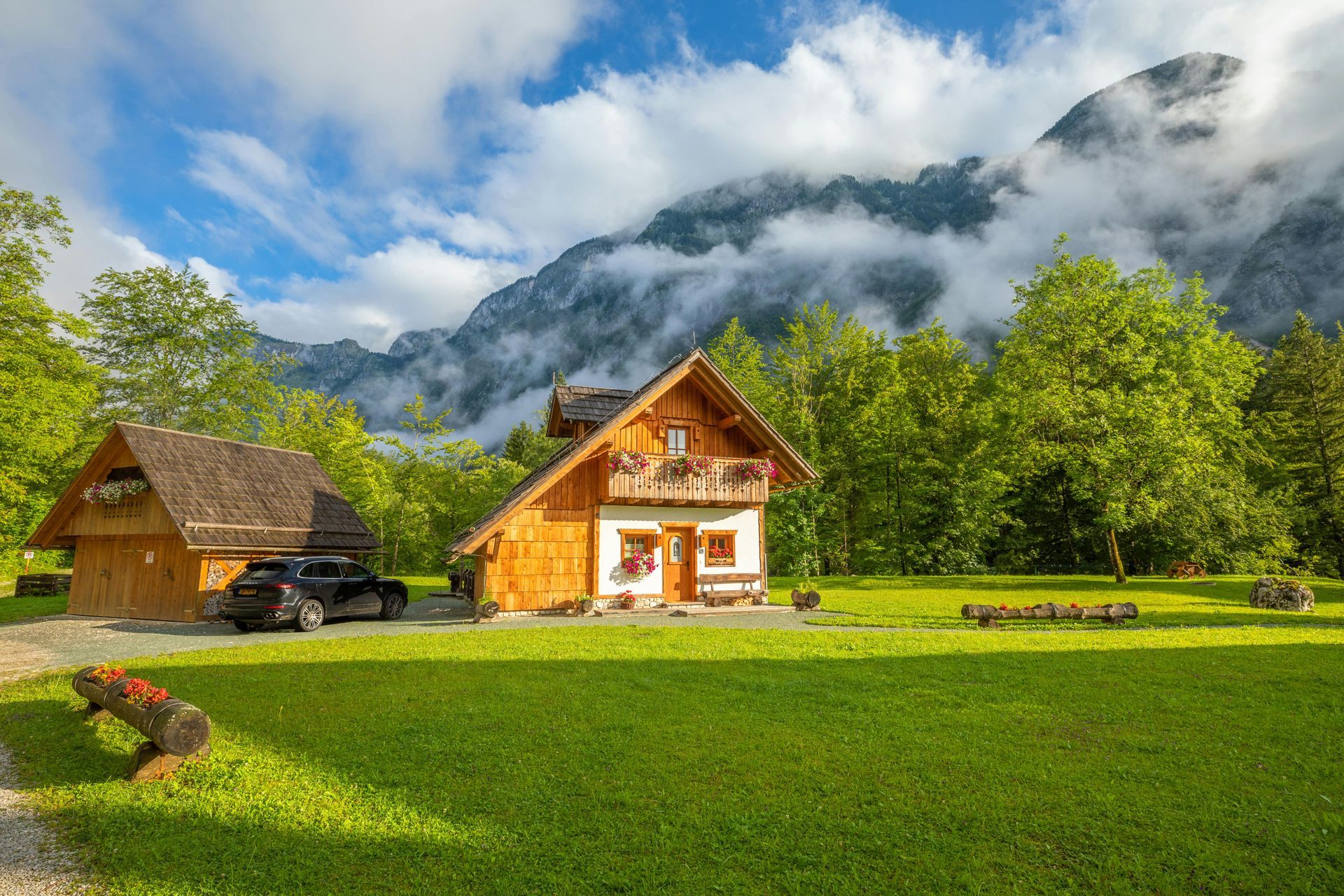Wooden chalet and outbuilding with a car in front, green lawn, mountains in background, cloudy sky.