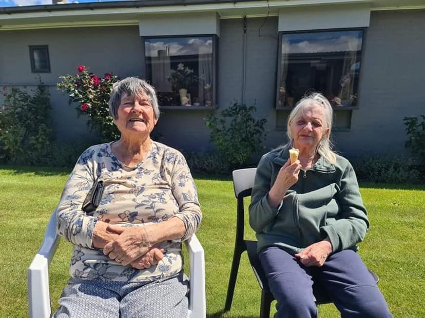 Two older women are sitting in chairs in front of a house eating ice cream.