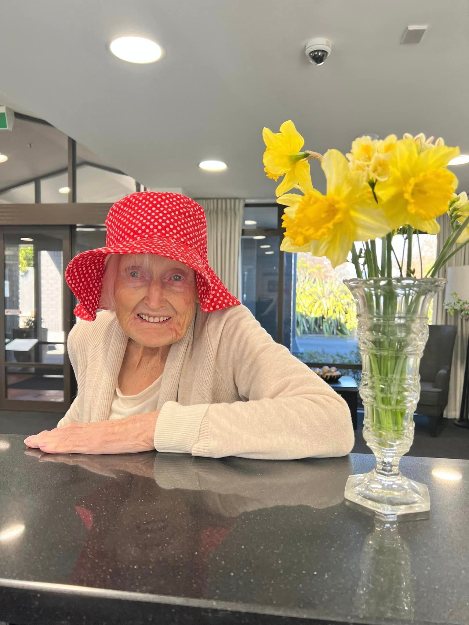 An elderly woman wearing a red hat is sitting at a counter next to a vase of yellow flowers.