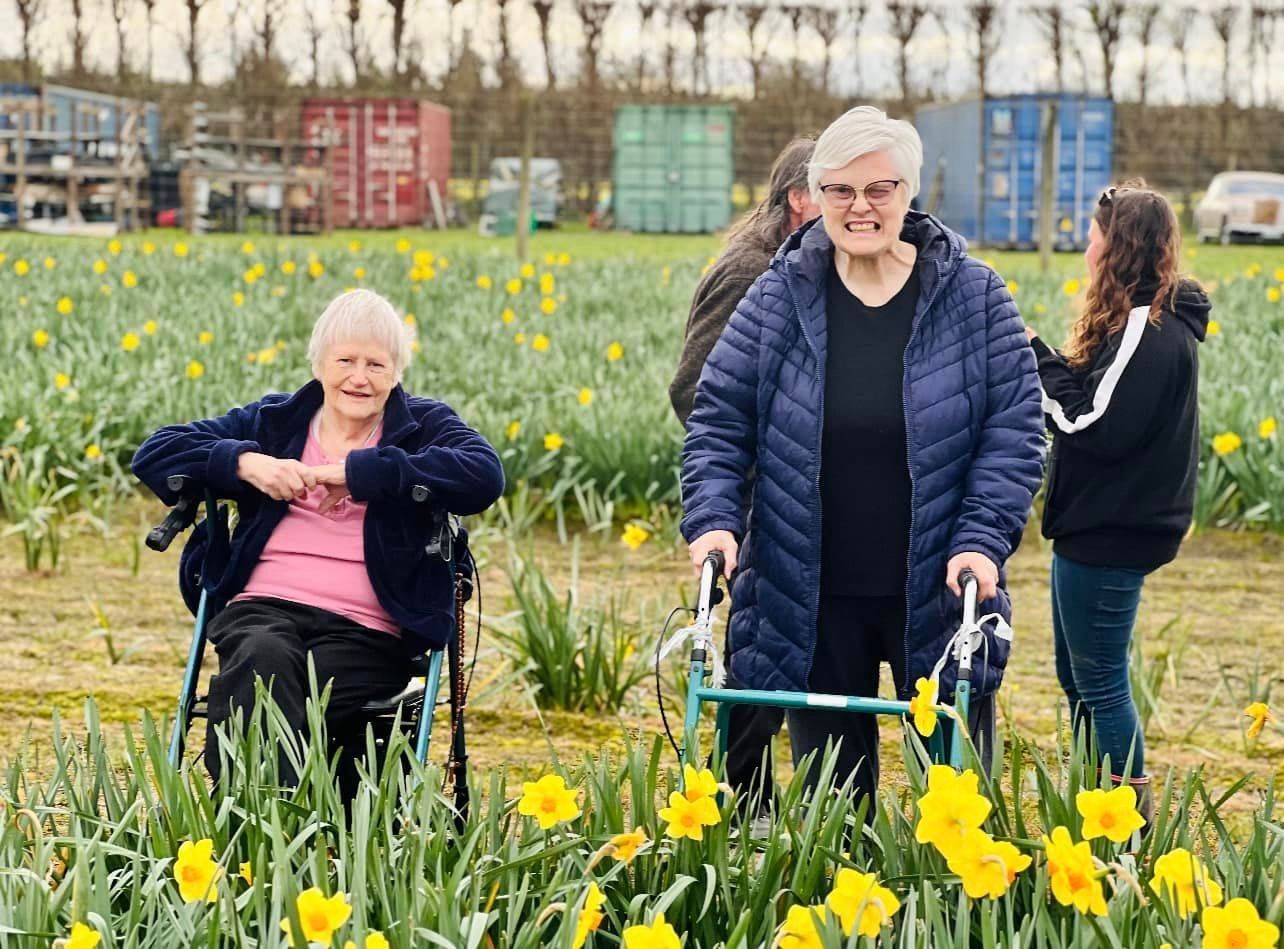 Two elderly women are standing in a field of yellow flowers.