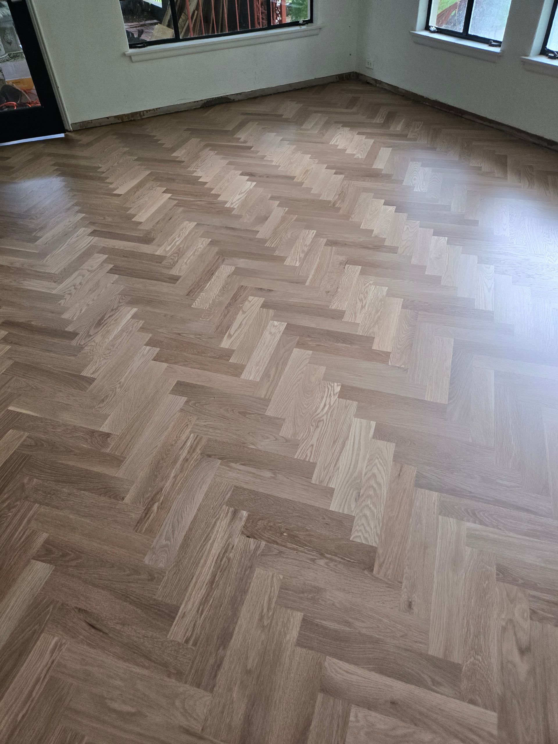Empty room with light wood herringbone flooring and windows along the walls.