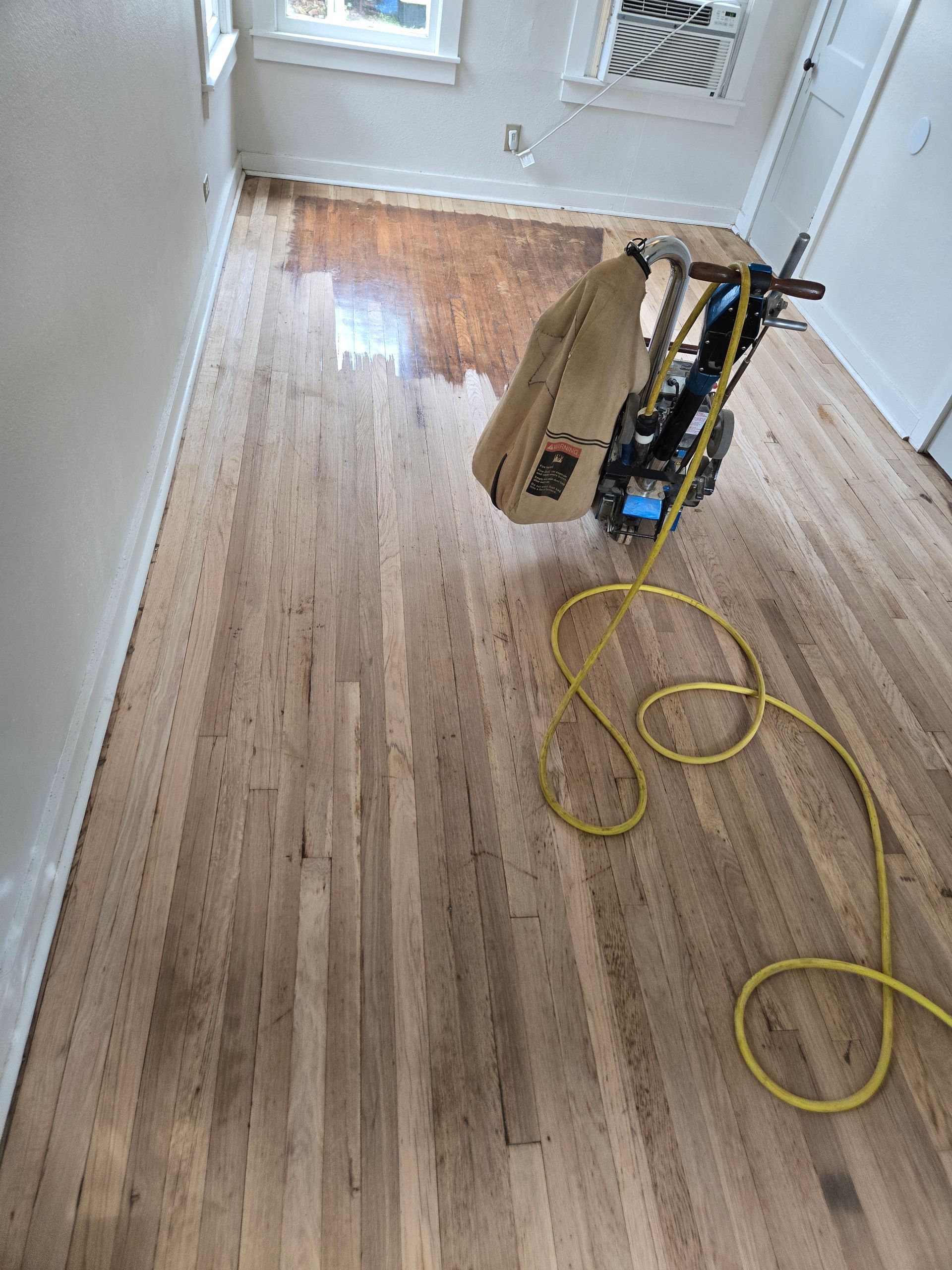 Hardwood floor refinishing in progress, with sanding equipment and yellow power cord in a hallway.