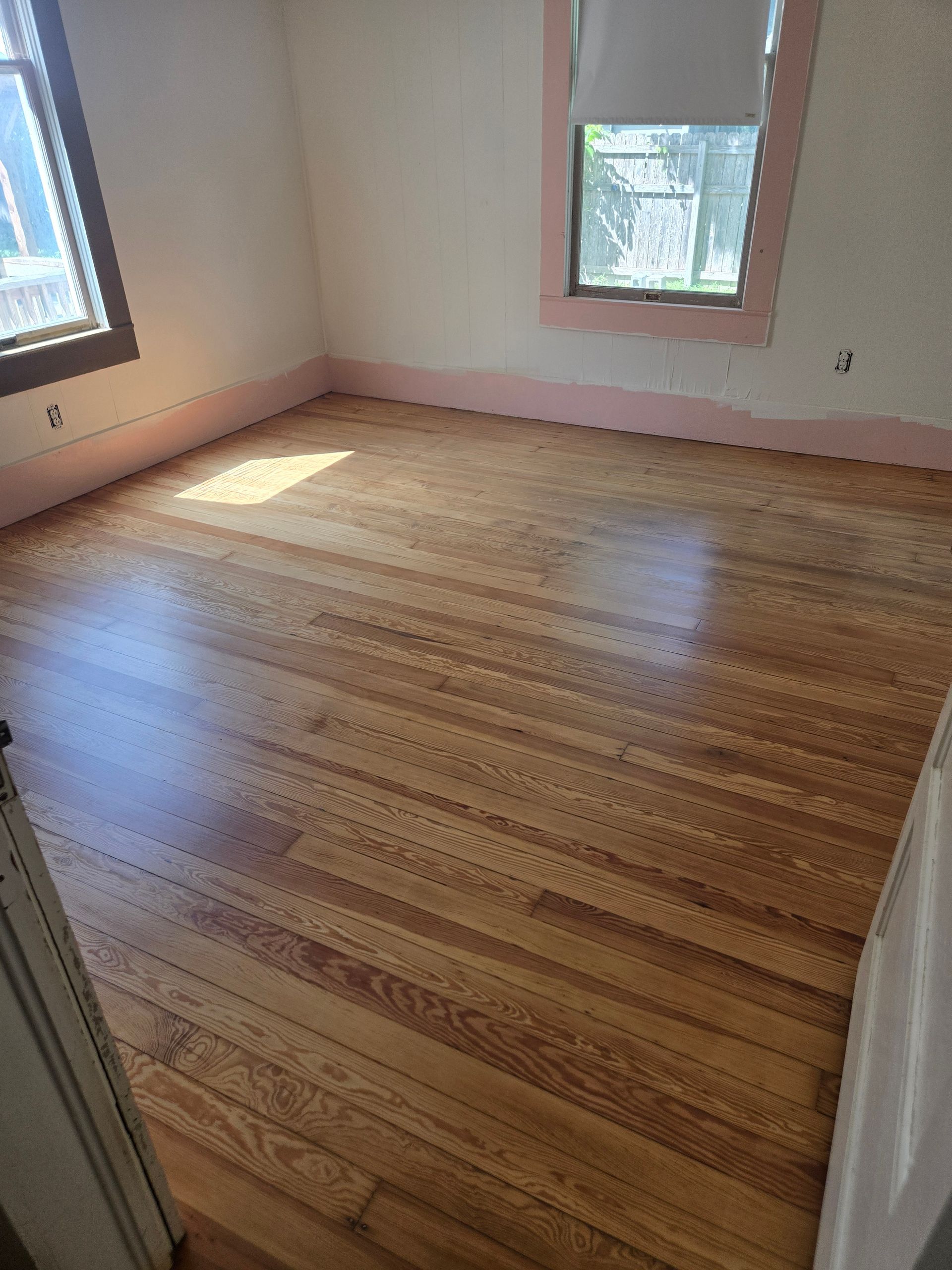 Empty room with hardwood floor, pale pink walls, and two windows letting in sunlight.