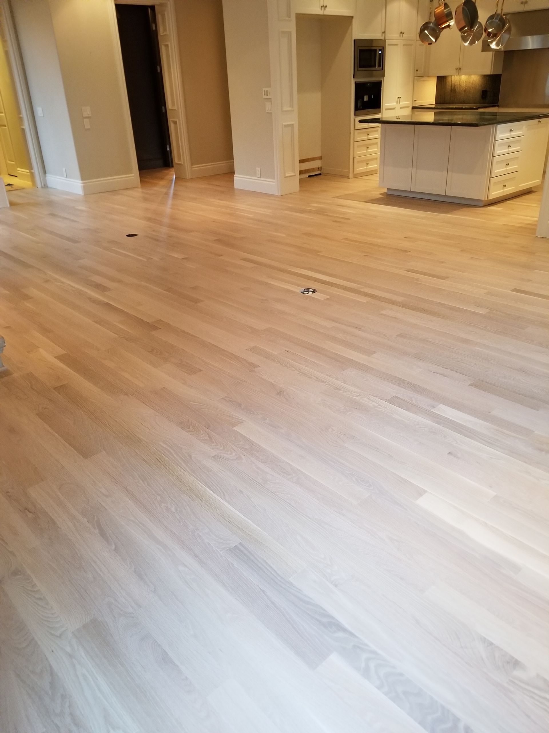 Empty hardwood-floored room with kitchen area and doorway in the background