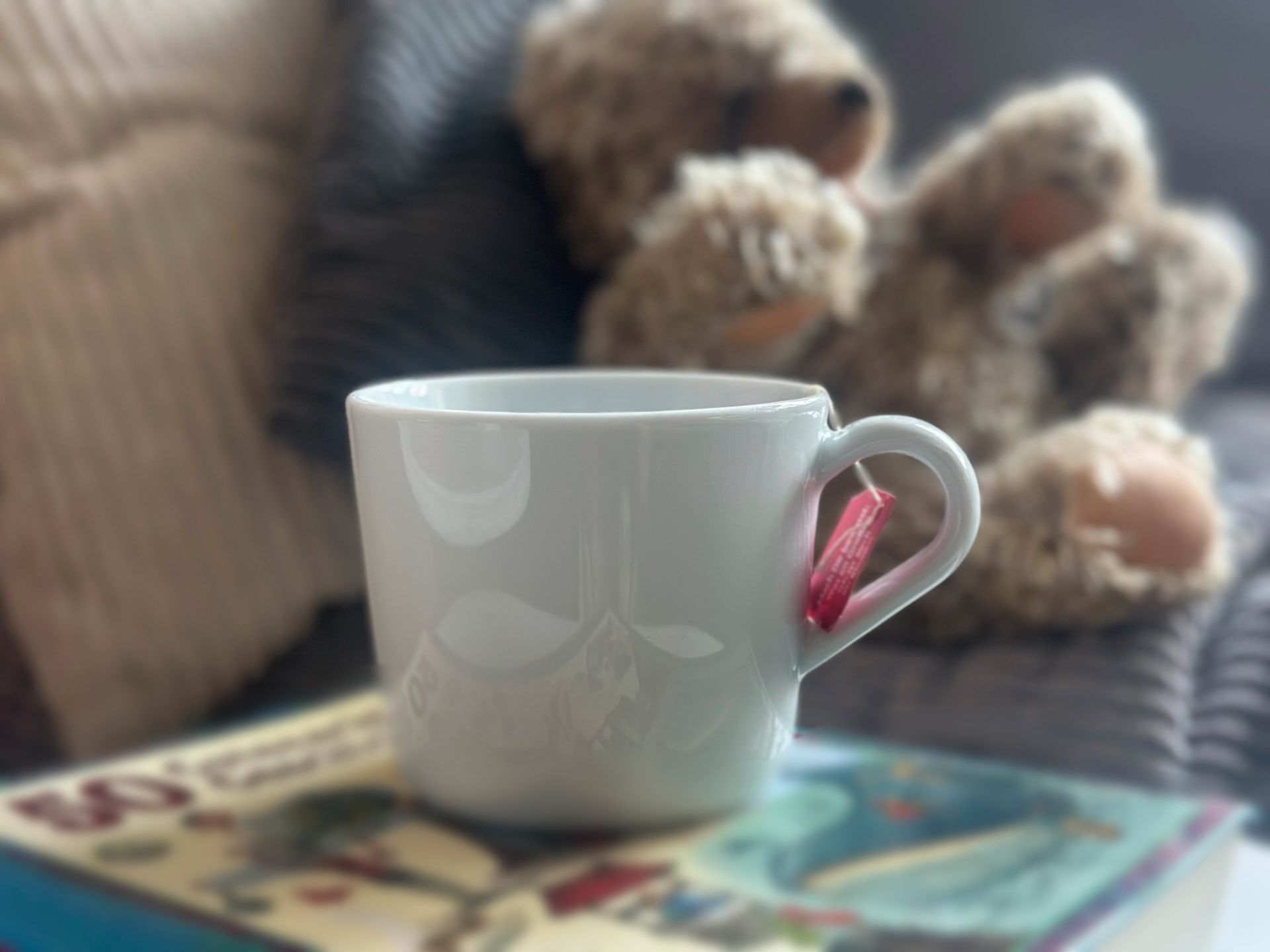 Weiße Tasse Tee auf einem Buch mit Gutenachtgeschichten, dahinter ein Teddybärauf dem Sofa