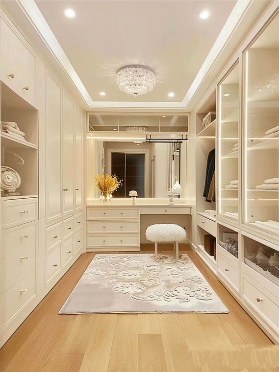 Elegant cream-colored walk-in closet with built-in shelves, vanity, chandelier, and rug on wood floor