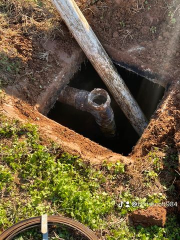 Open hole in ground with pipes, one large, rusty, and cut, and a white one resting across the opening.