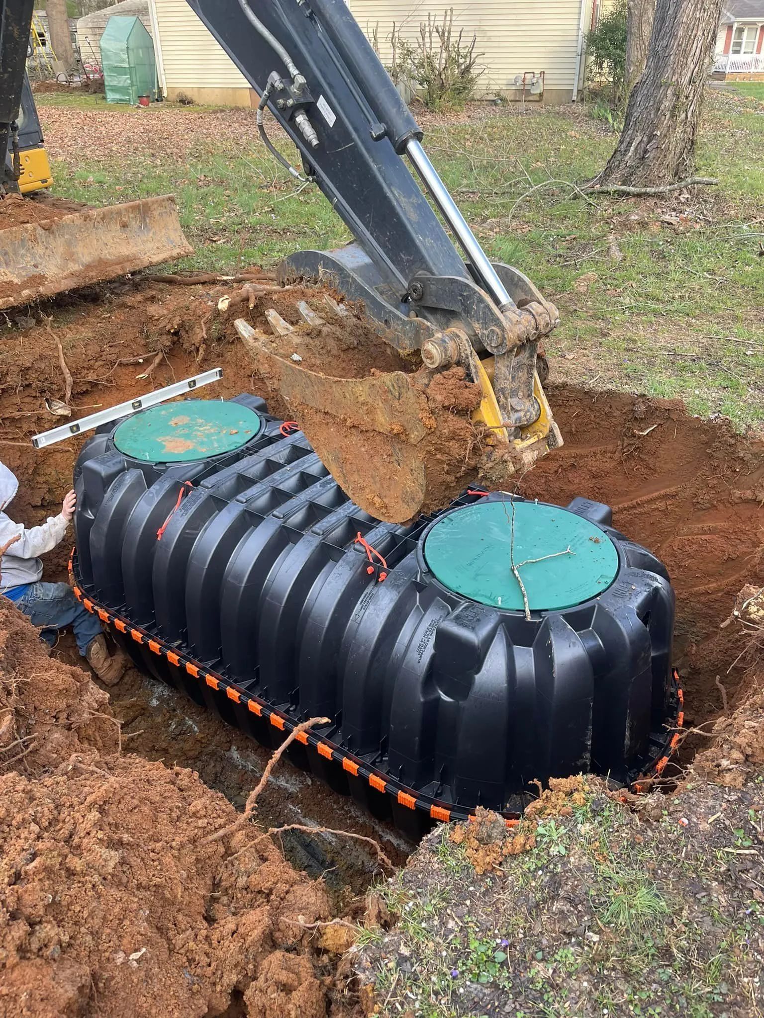 Backhoe installing a black underground septic tank with green caps in a dirt trench.