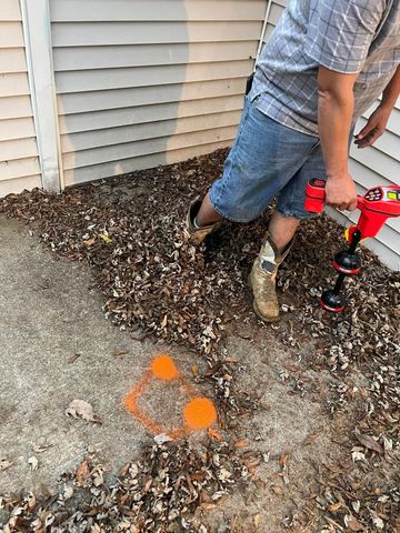 Person holds a red tool near orange markings on the ground outside a building, surrounded by leaves.