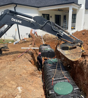 Excavator lowering a septic tank into a trench at a residential construction site.