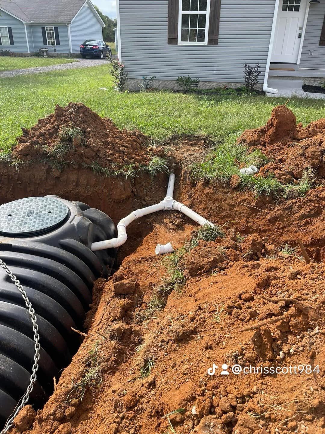 A black septic tank in an excavated area with white pipes leading away, near a house with gray siding.