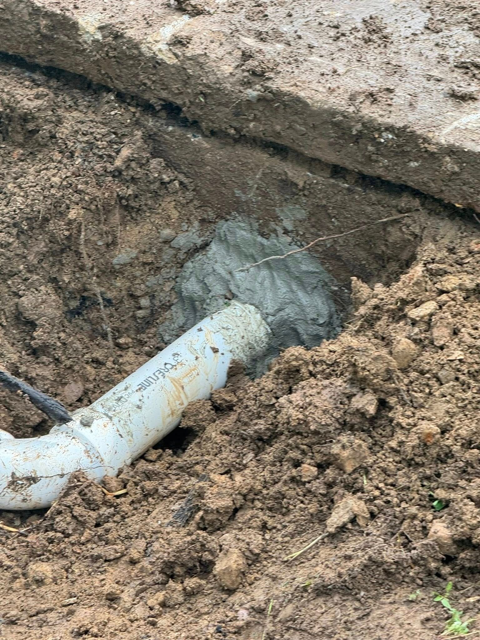White PVC pipe being joined in a muddy trench with wet concrete around the connection