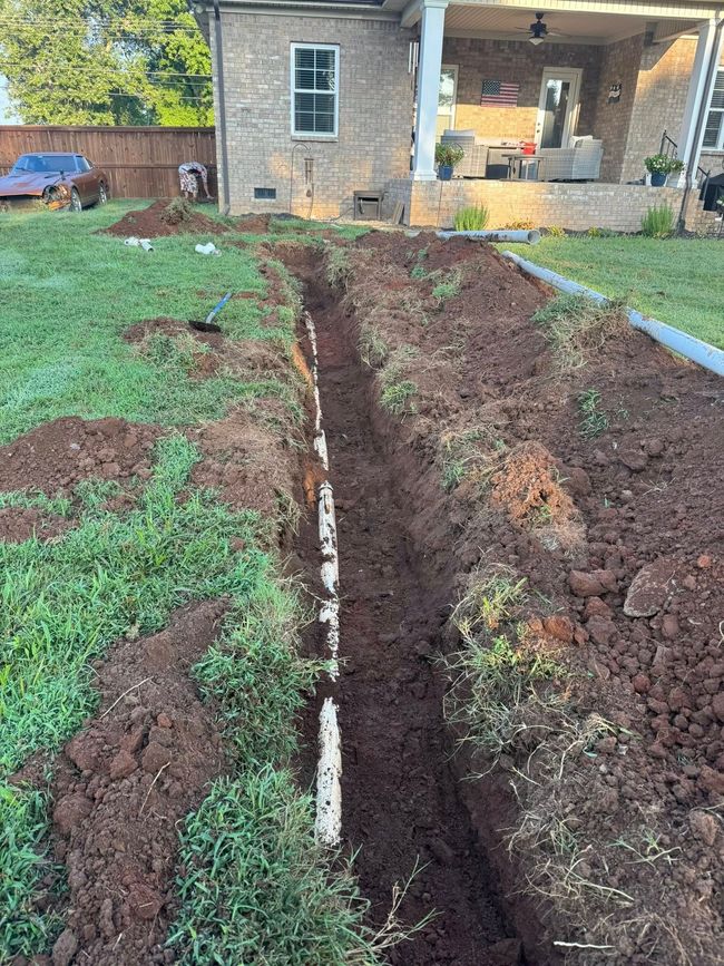 A trench dug in a yard with exposed white pipe, near a brick house with a porch.