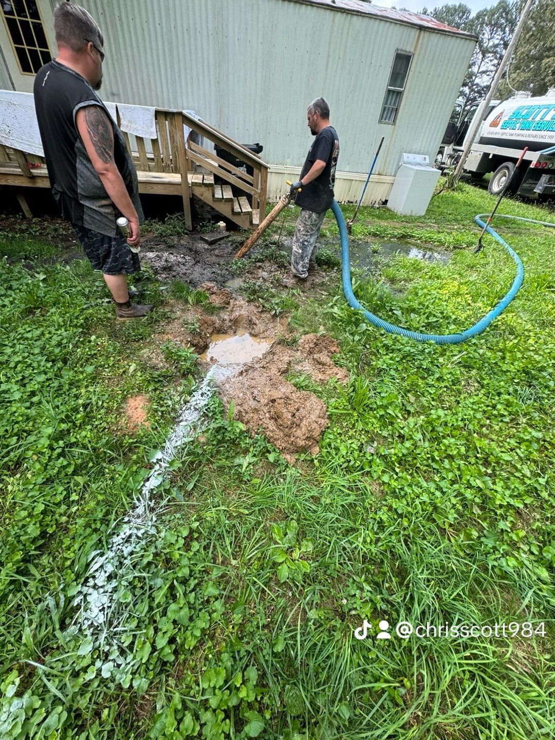 Two men outdoors cleaning up a muddy area near a house with a hose and shovel.