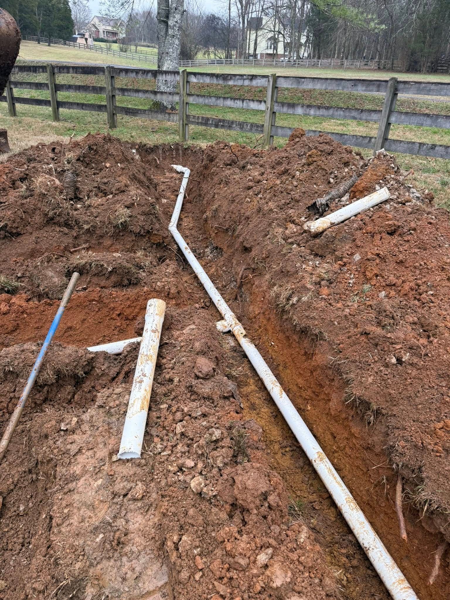 Trench with white pipes laid in it, surrounded by dirt. A shovel is also present. Outdoors.