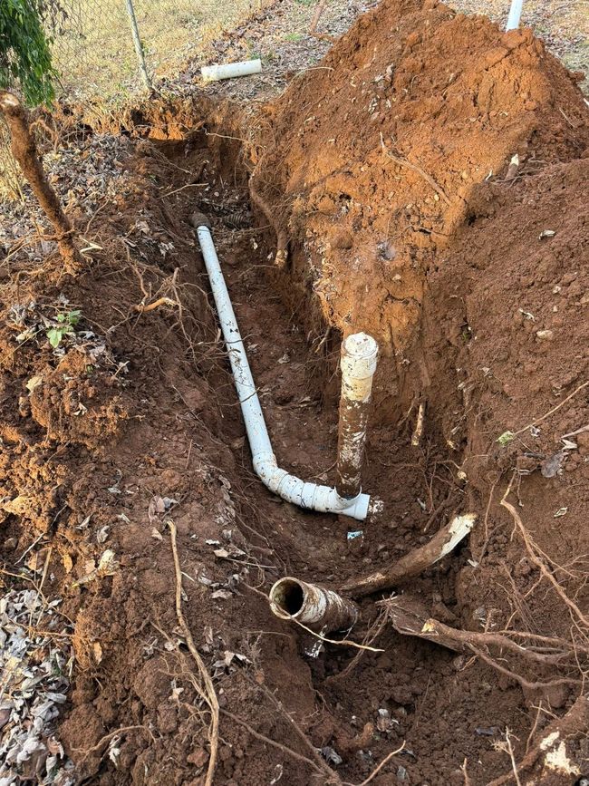 A trench reveals white PVC and copper pipes in dirt. Brown earth surrounds them.