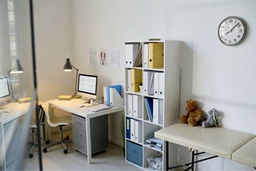 Small office with desk, computer, shelves, teddy bear, and wall clock near a window