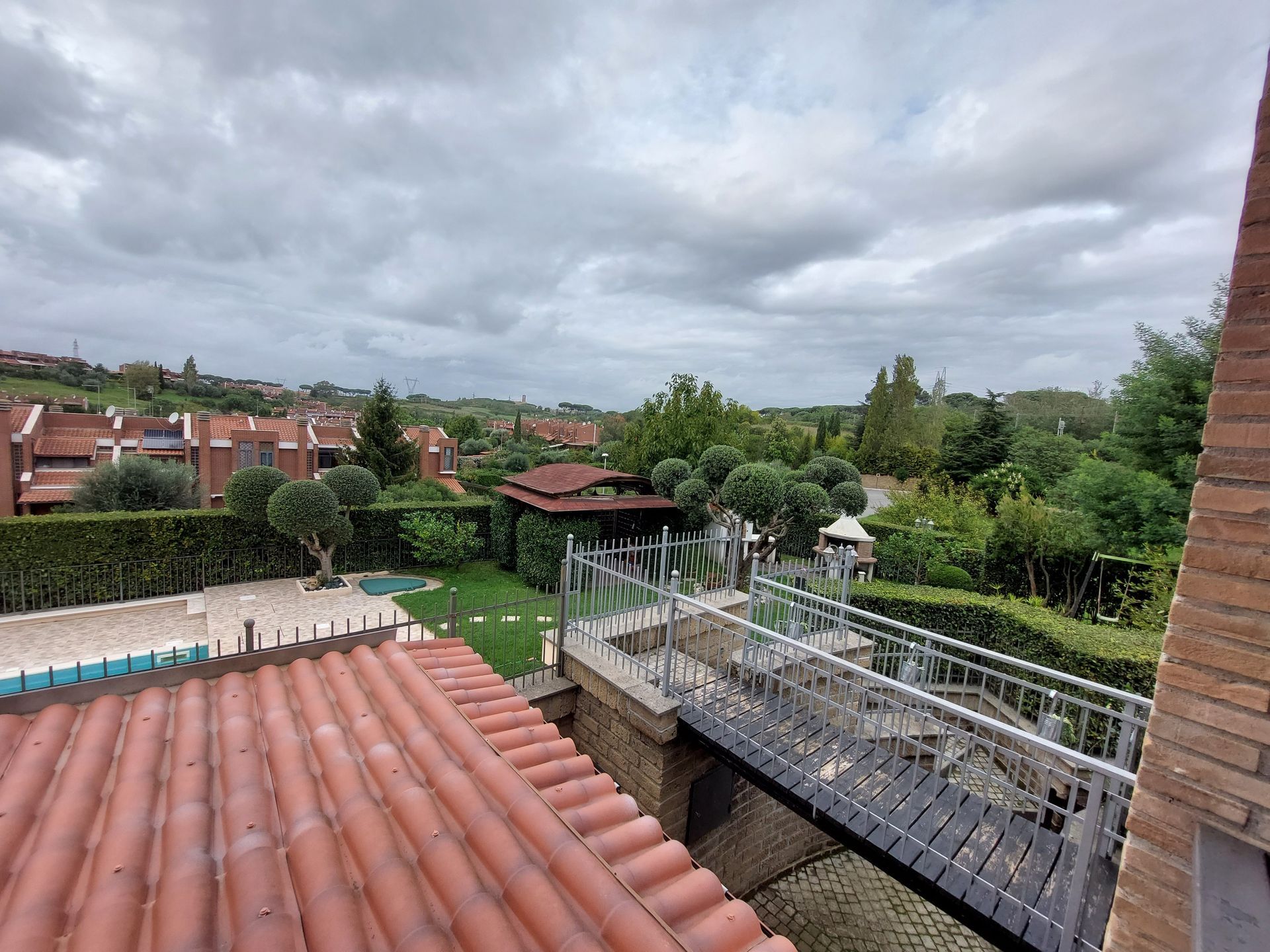 Vista da un tetto su tetti di tegole rosse, un giardino e un cielo nuvoloso.