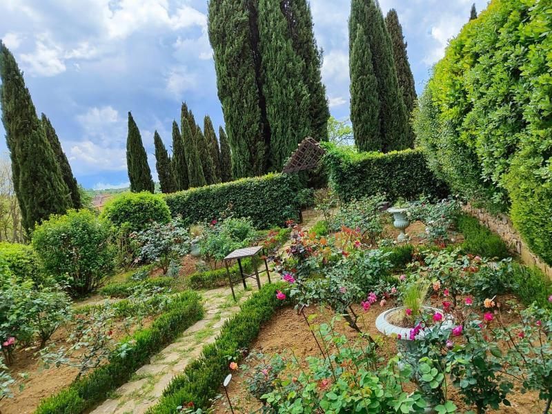 Un giardino lussureggiante con aiuole di rose, un sentiero di pietra e alti cipressi sotto un cielo parzialmente nuvoloso.