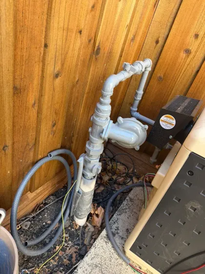 White PVC pipes and a pump beside a wooden wall, with a coiled gray hose on the ground.