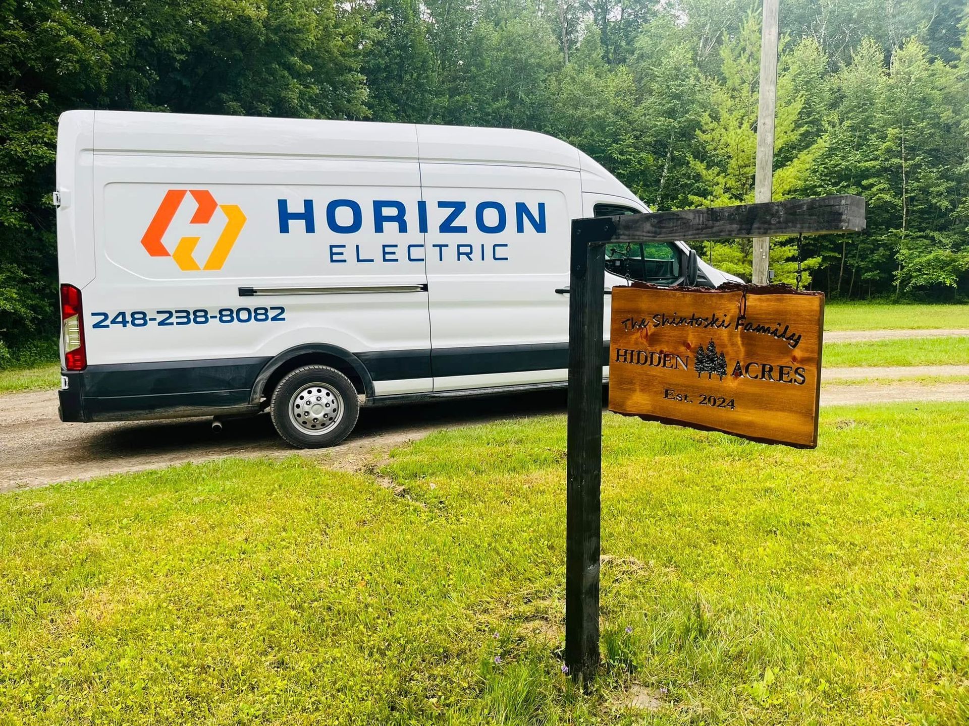 A white Horizon Electric service van is parked on a dirt road next to a hanging wooden sign on a metal post.