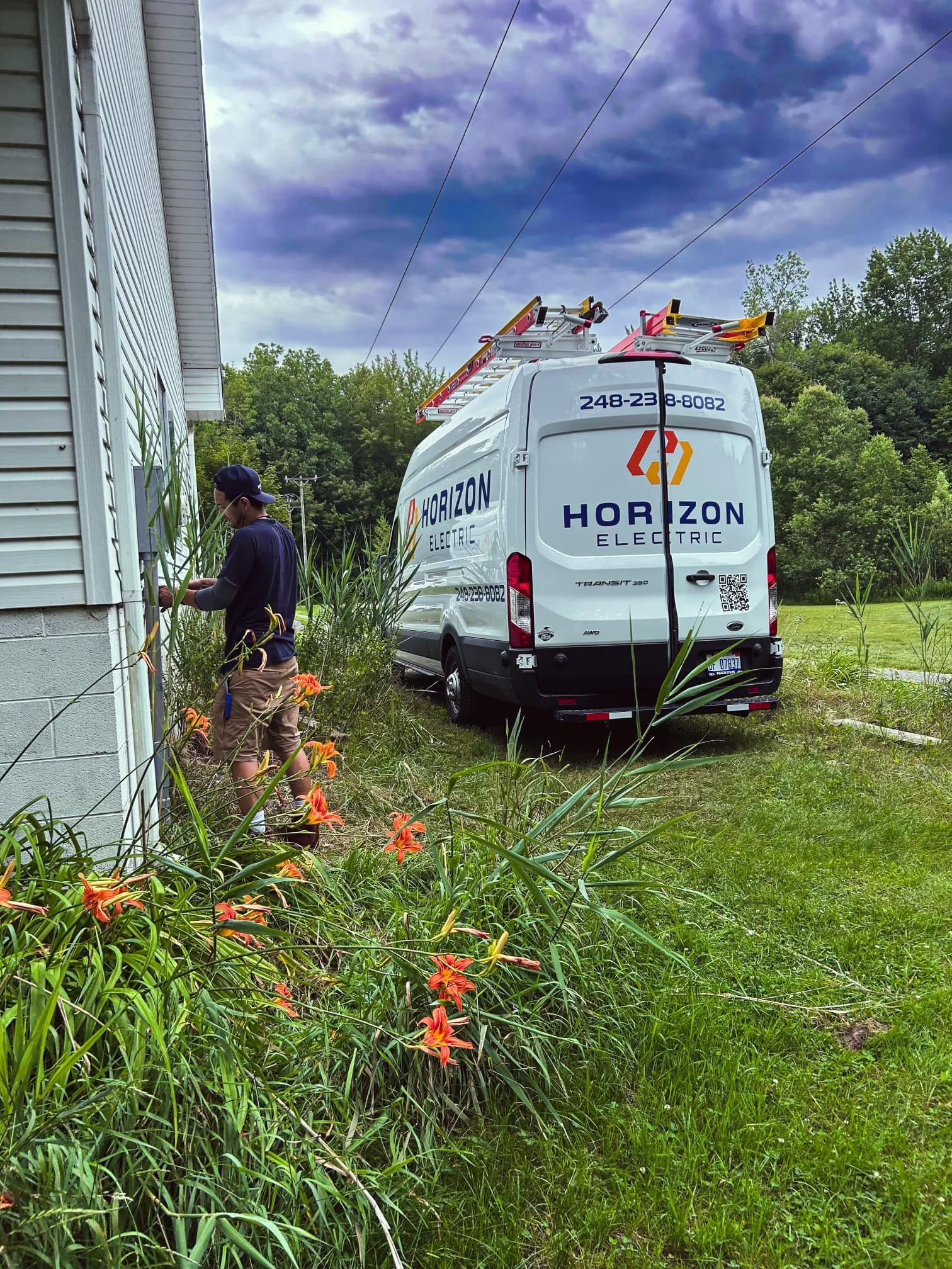 An electrician works on a home's exterior next to a white Horizon Electric service van parked in a grassy yard.