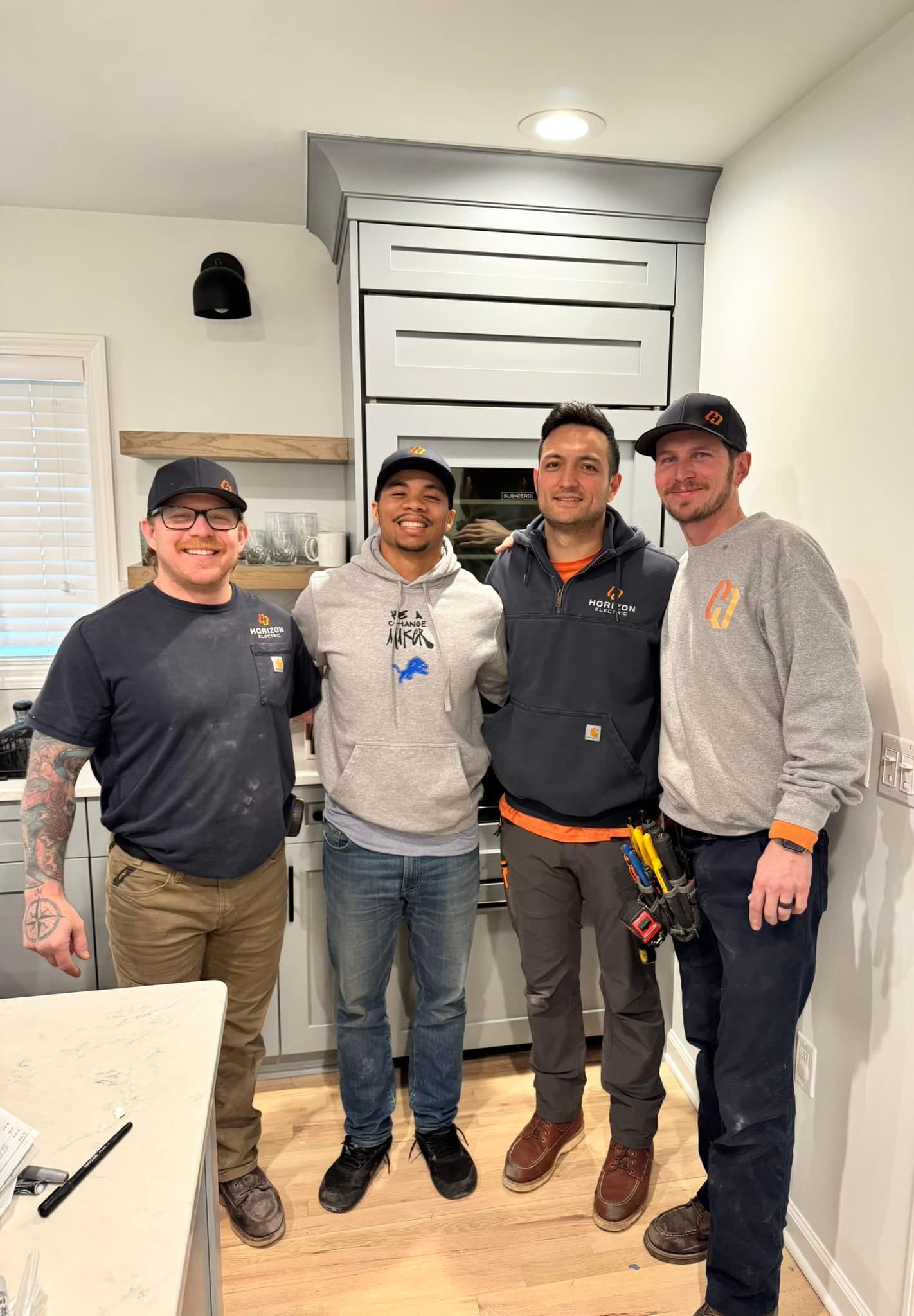 Four people in work attire stand side-by-side in a kitchen under construction, smiling at the camera.
