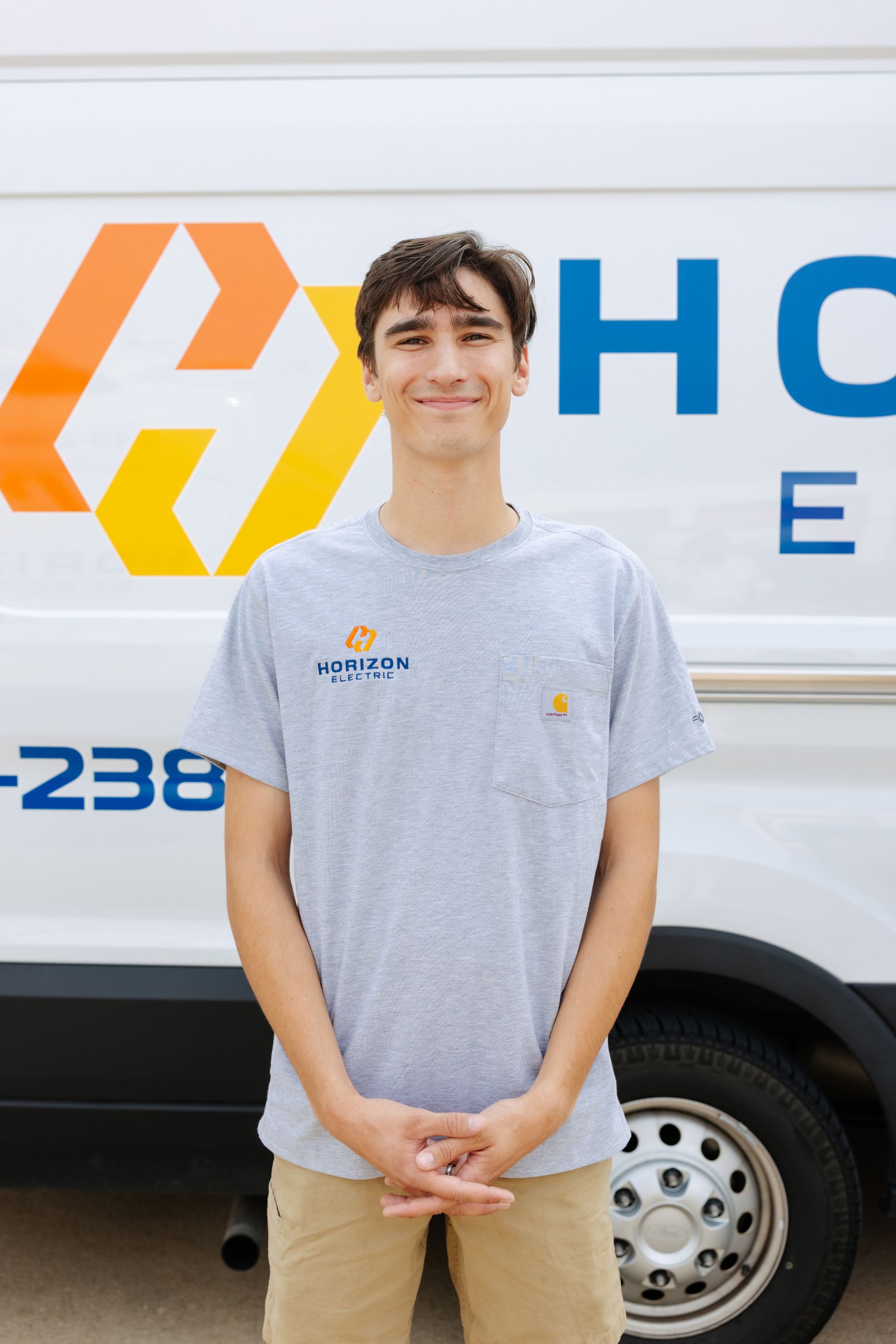 A smiling person wears a navy blue t-shirt with a pocket, standing in front of a red brick wall.