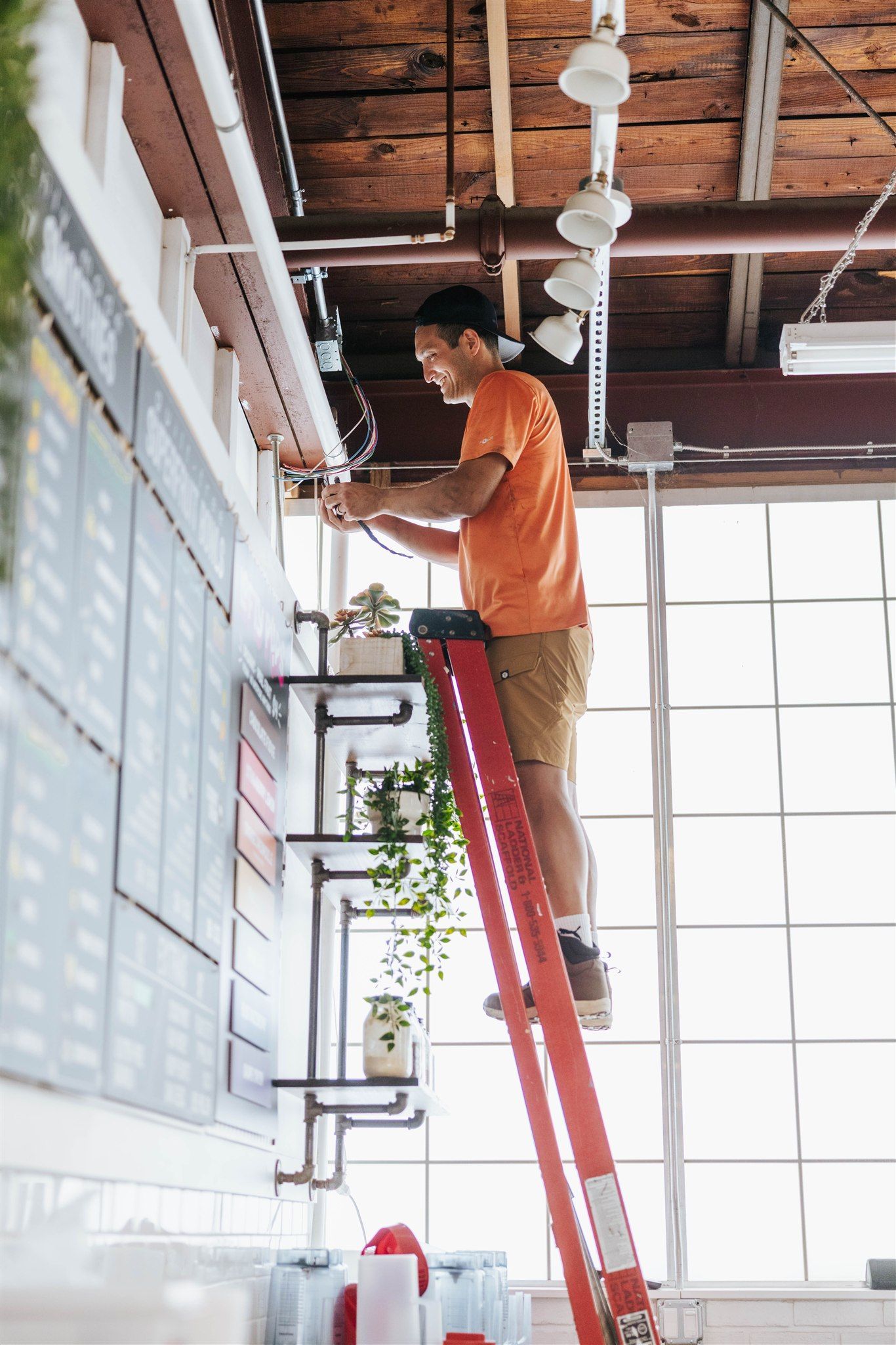 A person in an orange t-shirt and tan shorts stands on a red ladder, reaching up to work on light fixtures near a wall.