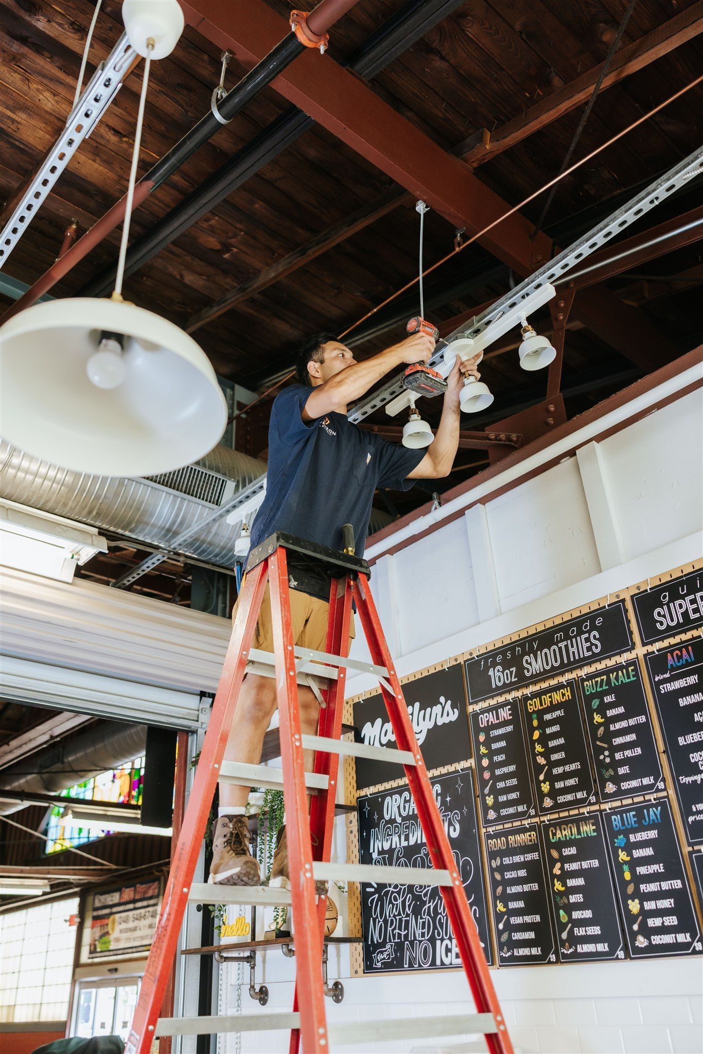 A person on a red stepladder works on overhead lighting fixtures in an industrial-style cafe with a chalkboard menu.