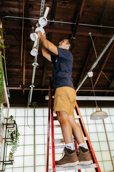 A person stands on a red ladder in a room with a rustic ceiling, performing maintenance work on the overhead lights.