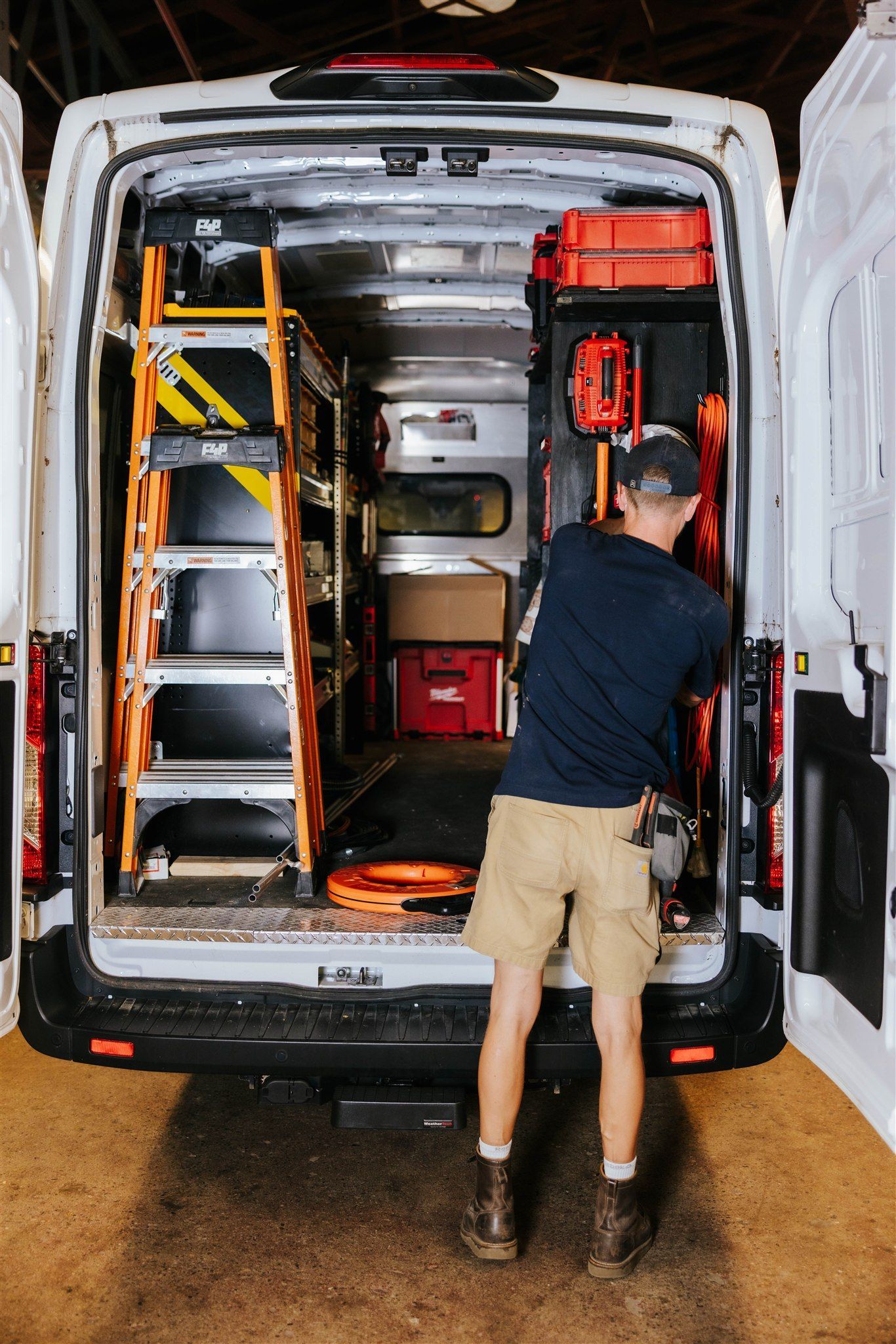 A worker in a dark shirt and khaki shorts organizes tools and supplies inside the back of a white utility work van.
