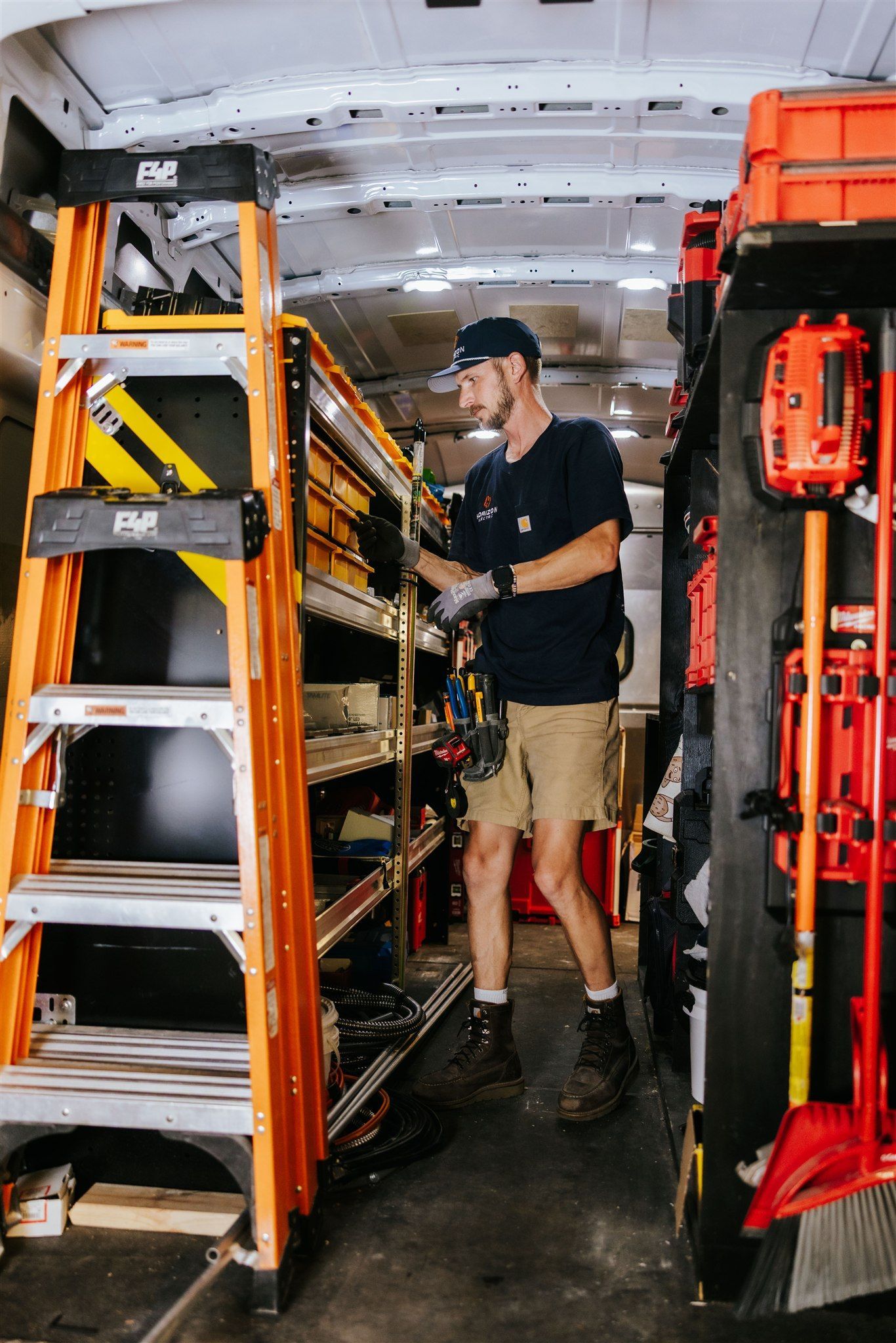 A technician standing inside a work van, organizing tools on metal shelving units next to an orange folding ladder.