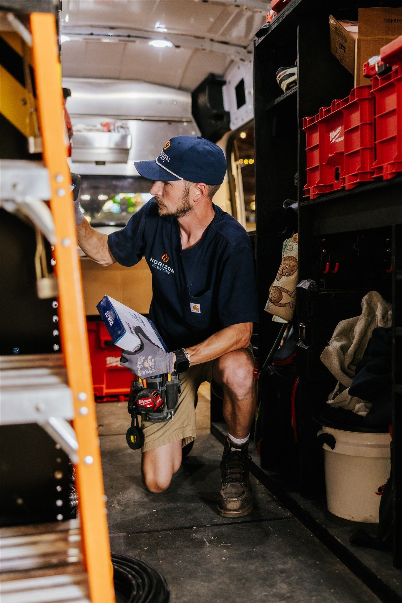 A person wearing a blue cap and work shirt kneels inside a van, organizing tools and supplies on shelving units.