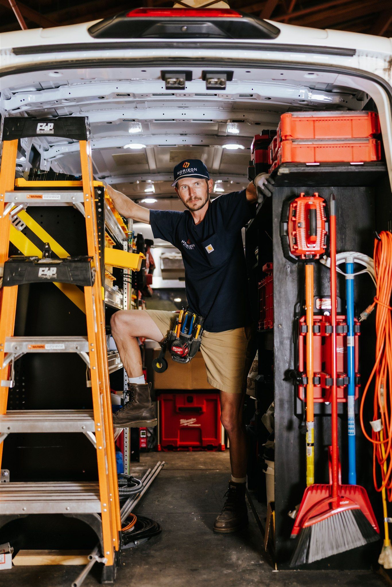 A person stands inside a work van, organized with ladders, tools, and equipment, looking toward the camera.