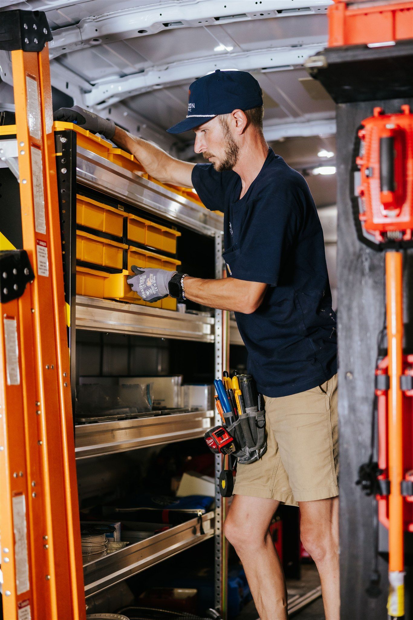 A technician in a navy shirt and cap organizes components in yellow bins inside a utility van filled with equipment.