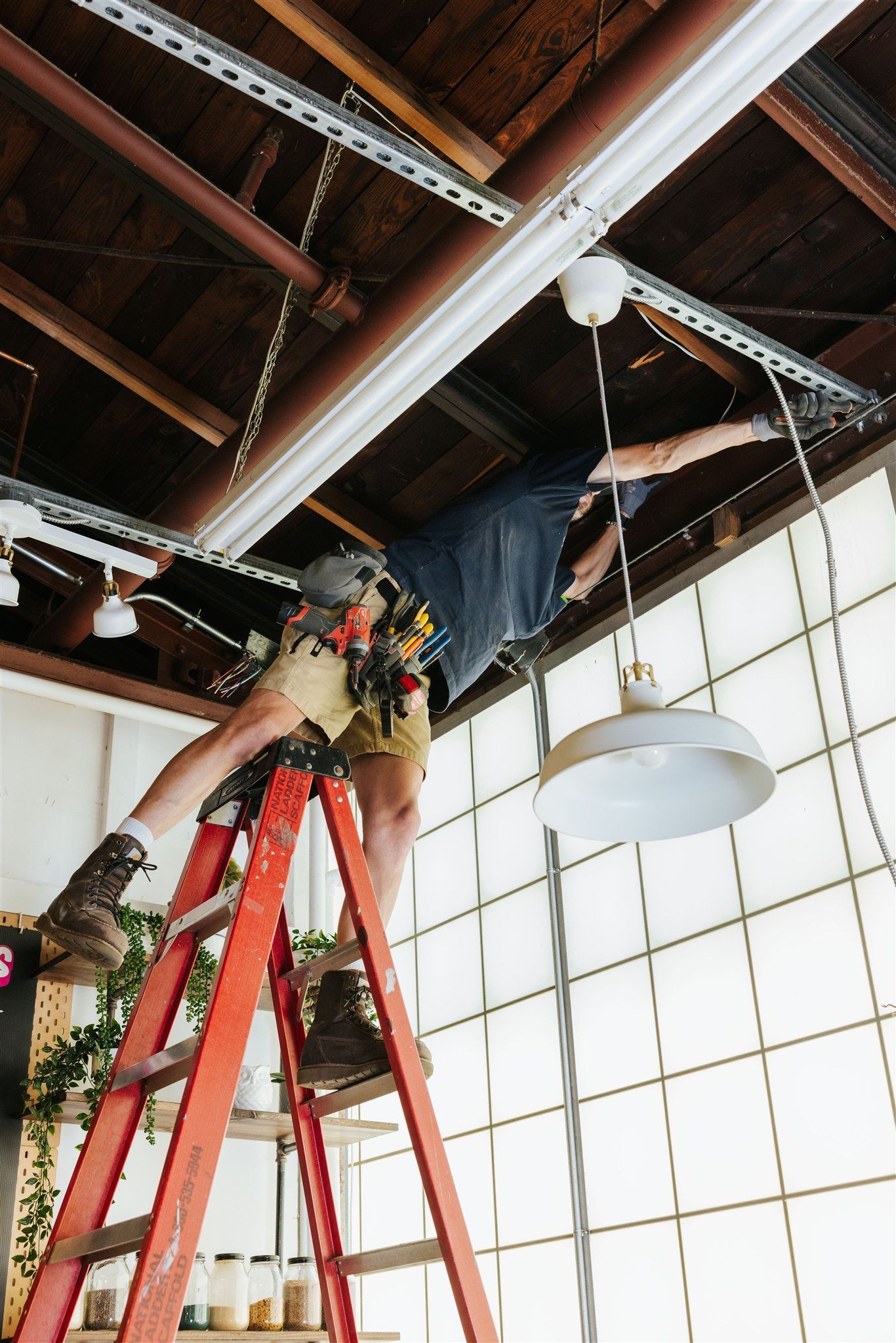 A person wearing a tool belt stands on a red ladder, working on a light fixture attached to an exposed ceiling joist.