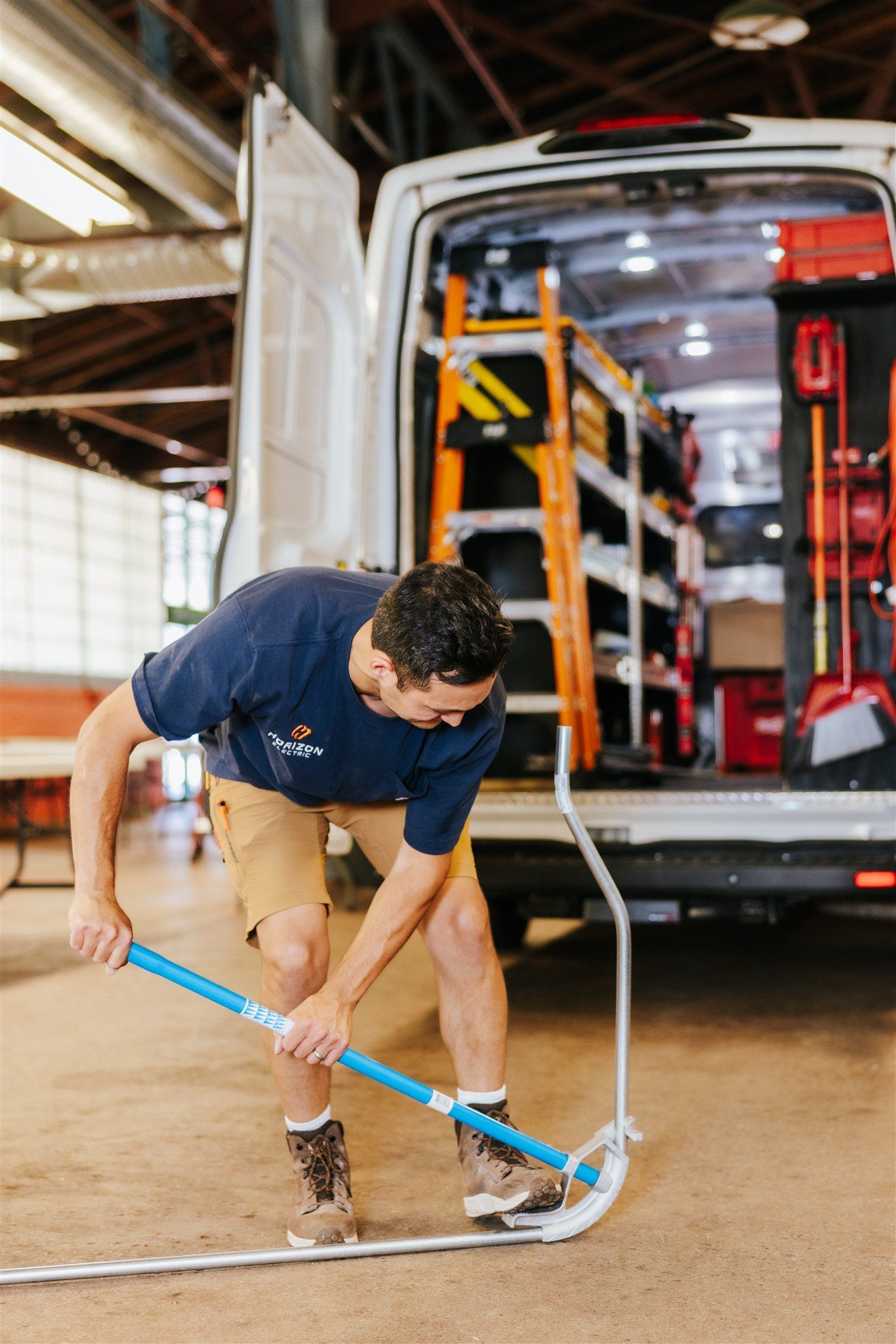 A worker wearing a blue shirt and khaki shorts uses a blue manual tool to install a metal frame on a concrete floor.