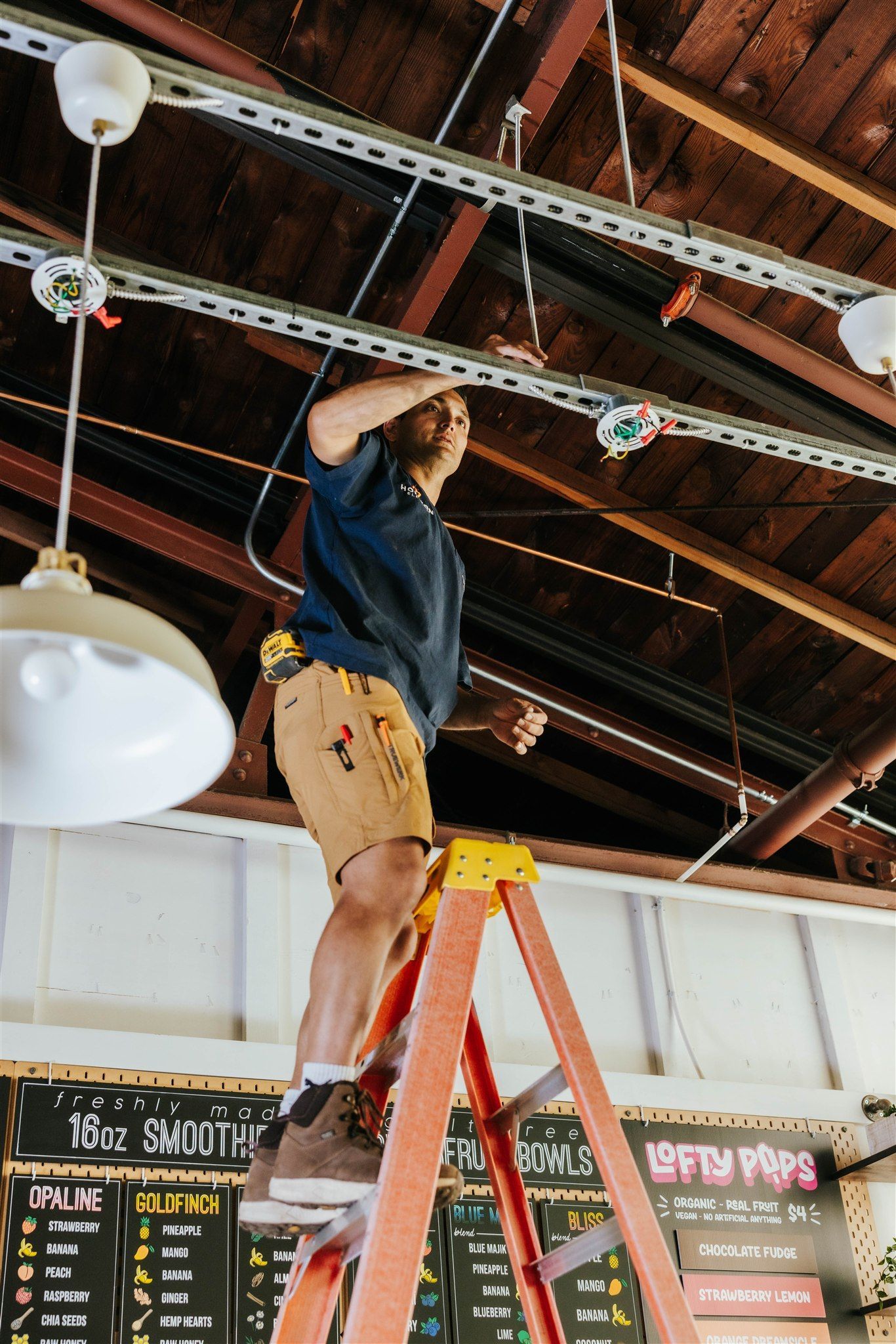 A person stands on a tall ladder, reaching up to work on electrical conduit mounted to a wooden ceiling in a workspace.