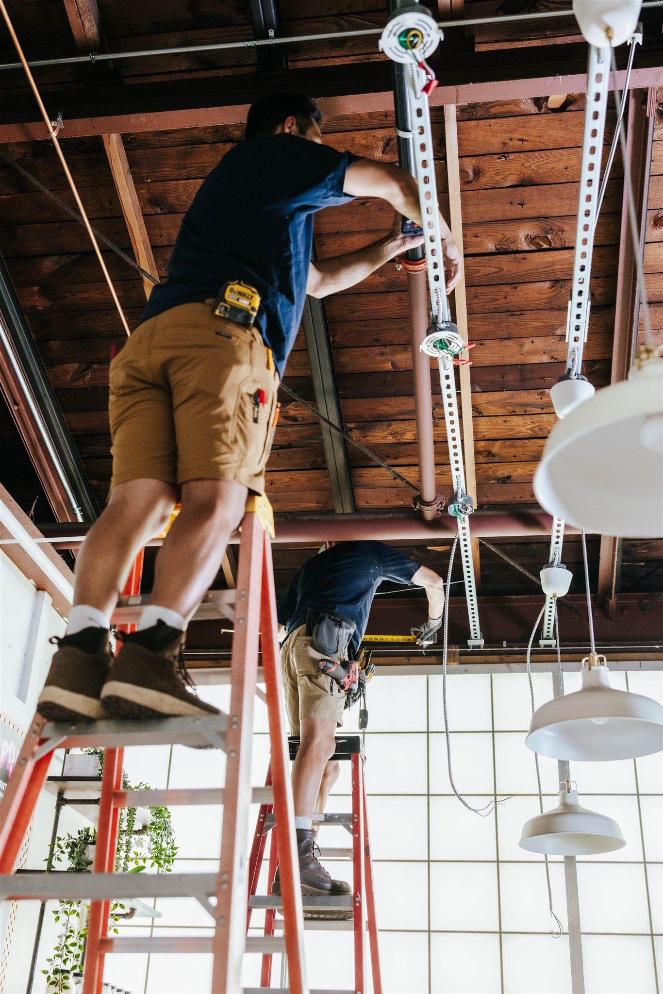 Two workers stand on ladders installing light fixtures from the wooden ceiling beams of an industrial-style workspace.